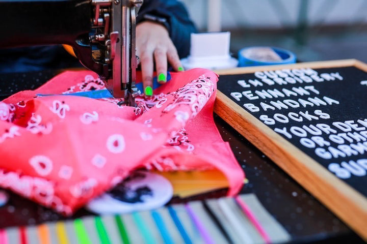 Photo credit: @100days.denver. Someone with green fingernails is live chainstitch embroidering a bandana. There is also a thread color card and a letter board with the embroidery options, including bandanas, socks, pennants, and more.